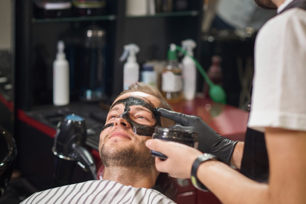 Young smiling male lying on chair in barber shop while barber in protective gloves putting black mask against black dots on face of client. Concept of cleaning procedure and face care.