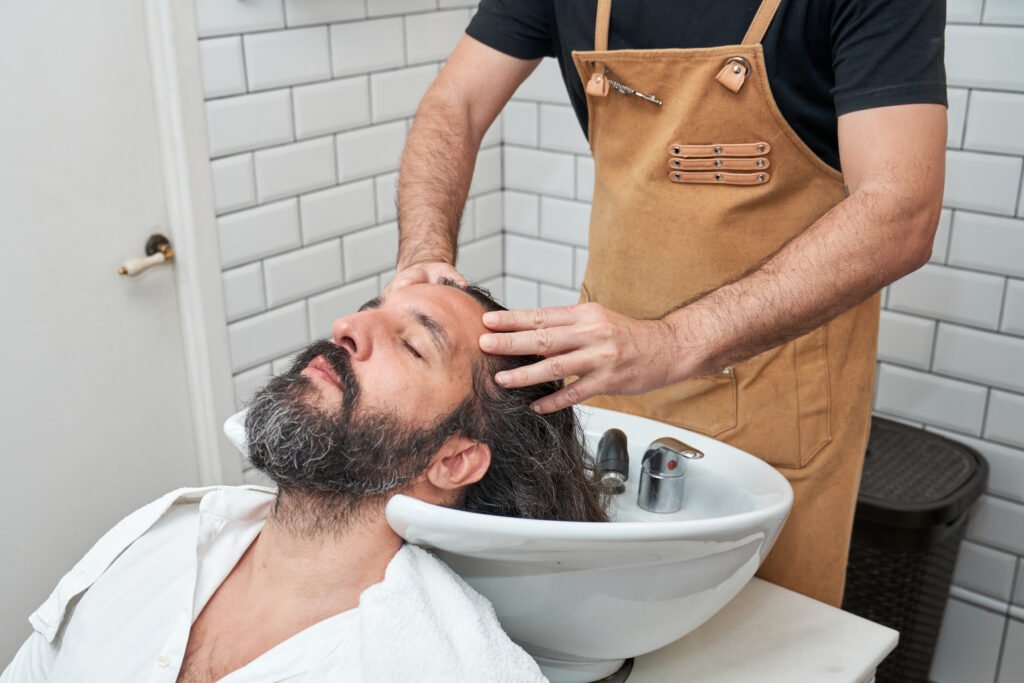 Anonymous barber in apron washing long hair of man with shampoo in white sink in professional barbershop